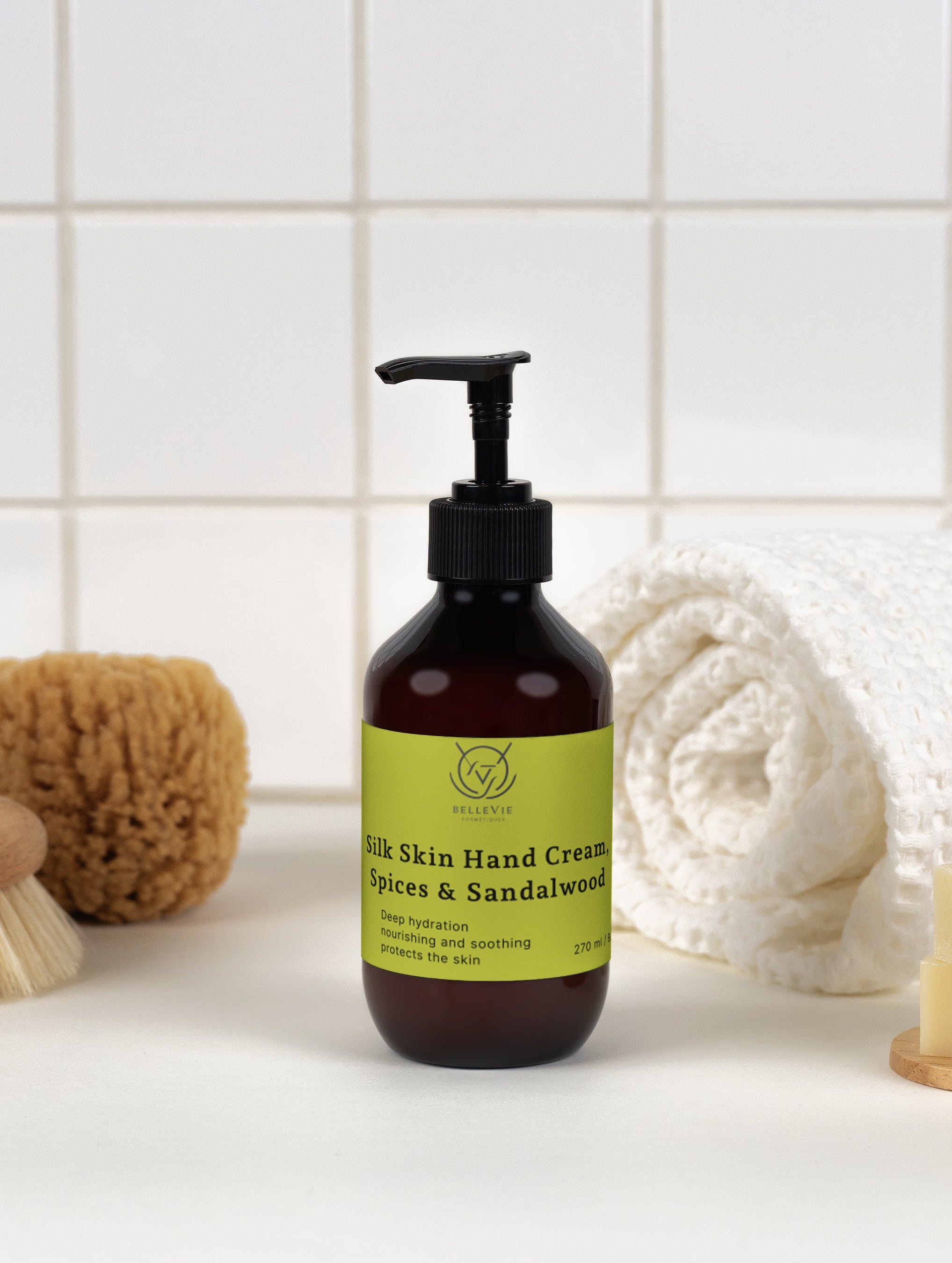 A dark amber pump bottle of Hand cream with a black dispenser and a lime green label, styled on a white surface with a white tiled background and bath accessories including a towel, soap, and brushes.
