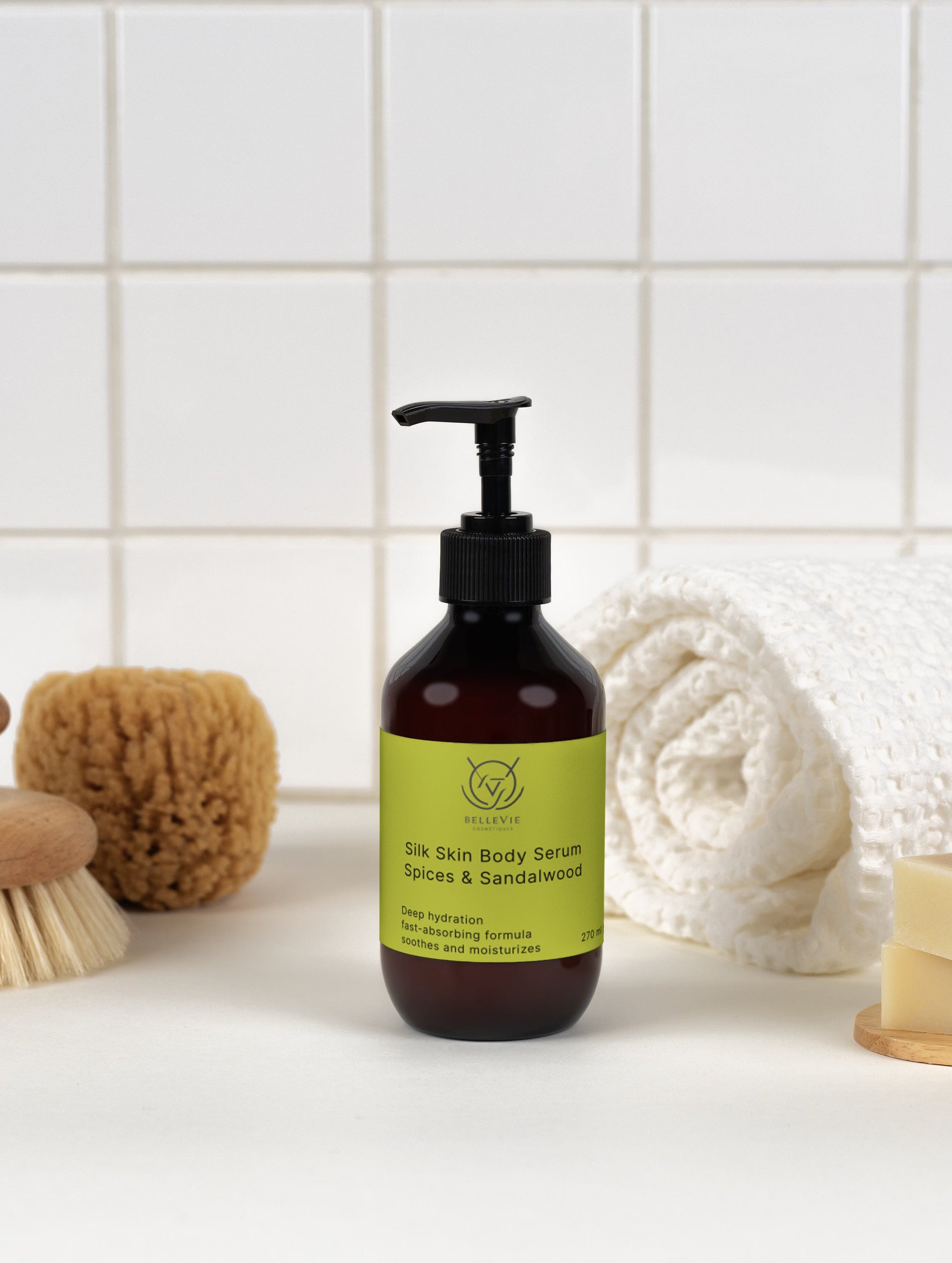A dark amber pump bottle of Silk Skin Body Serum with a black dispenser and a lime green label, styled on a white surface with a white tiled background and bath accessories including a towel, soap, and brushes.